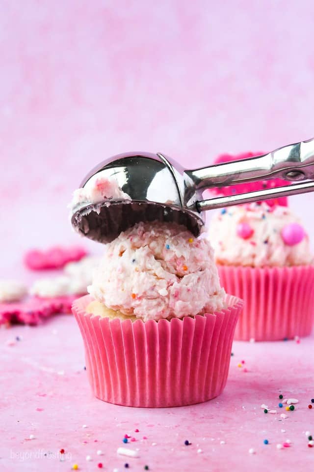 A cookie scoop placing frosting on top of a cupcake with a pink wrapper