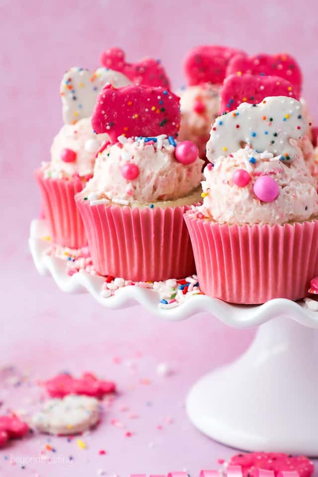 A white cupcake stand filled with cupcakes that have pink wrappers and are frosted and decorated with pink and white Circus Animal Cookies