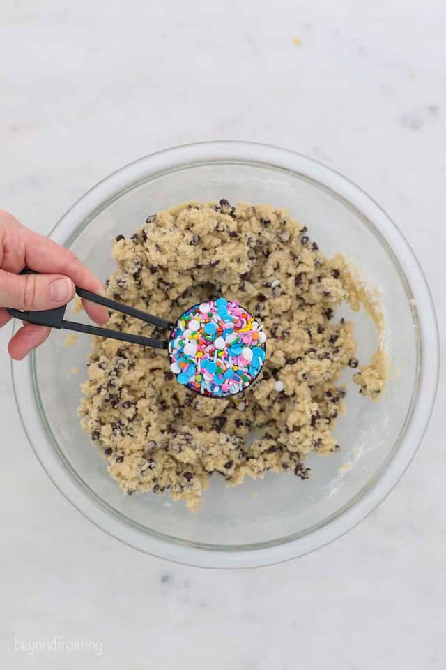 a hand holding a small measuring cup filled with sprinkles, hanging over a glass mixing bowl of cookie dough