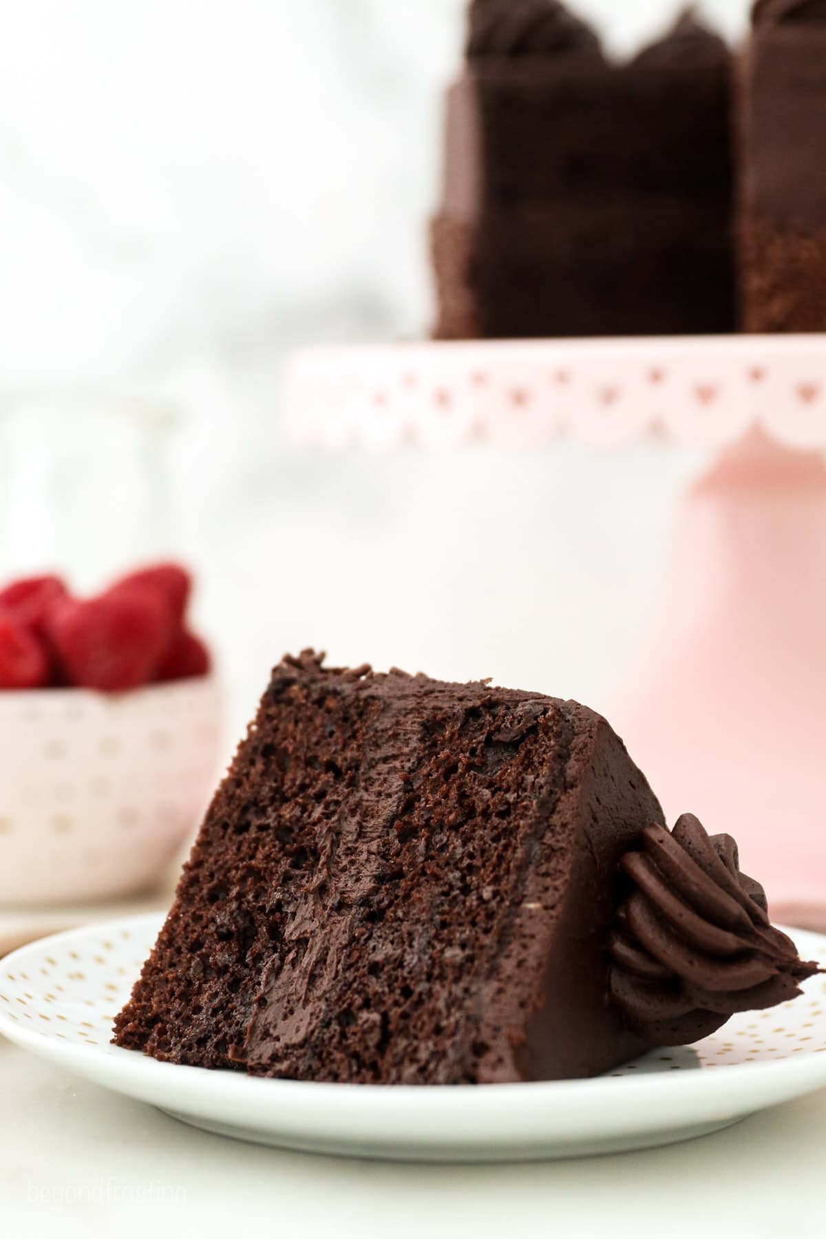 A slice of small chocolate cake topped with a swirl of chocolate buttercream laying on its side on a white plate, with the rest of the cake on a pink cake stand in the background.