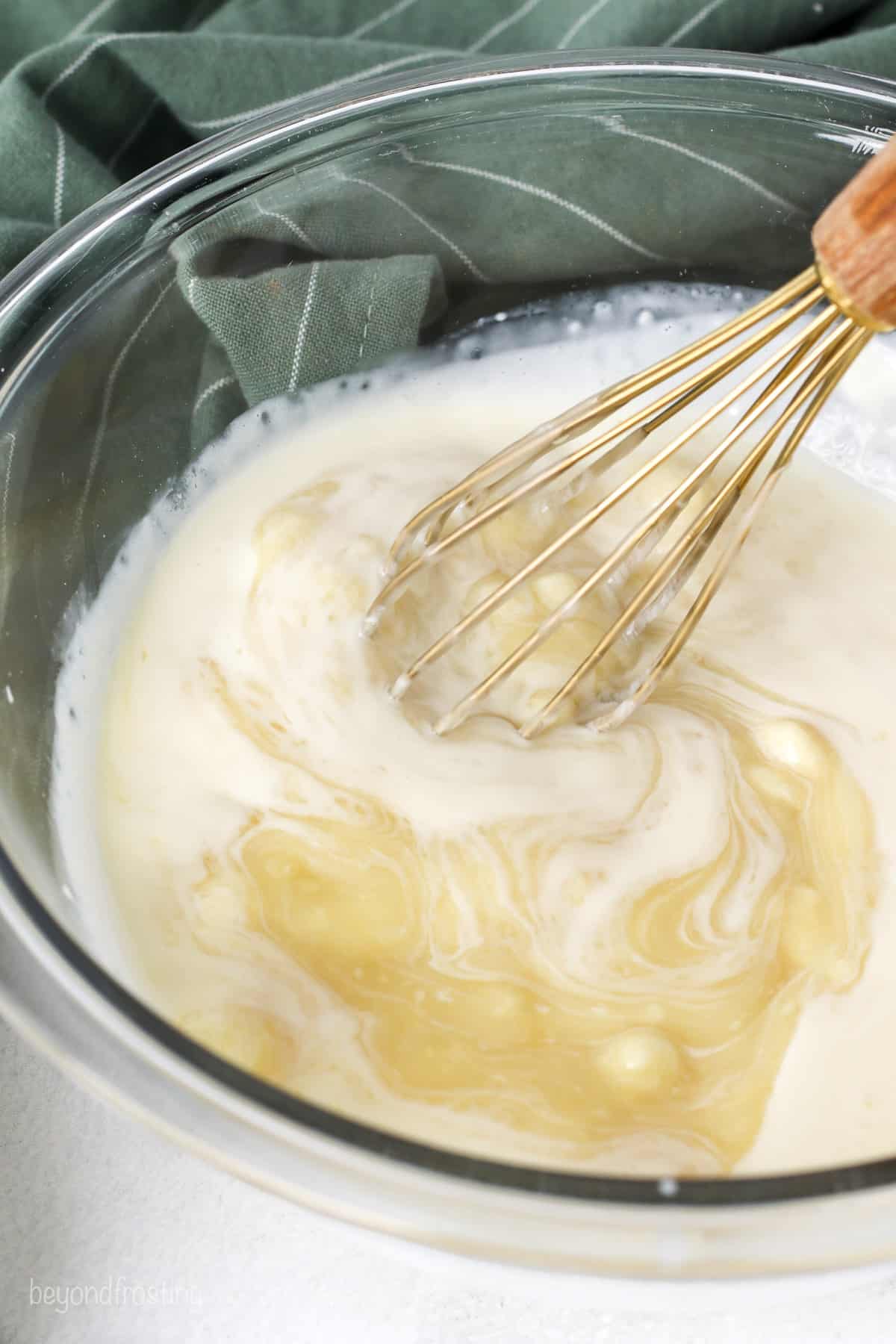 Melted white chocolate and heavy cream are whisked together in a glass bowl.