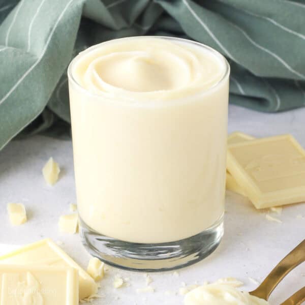 A glass of white chocolate ganache next to scattered white chocolate pieces.