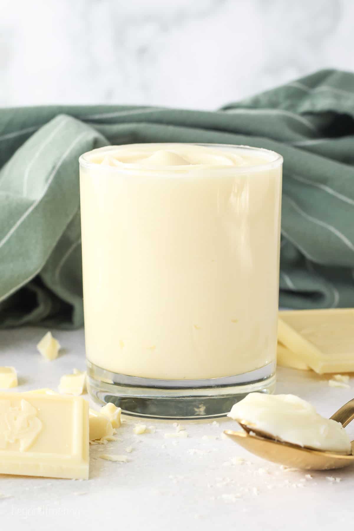 A glass of white chocolate ganache next to scattered white chocolate pieces.