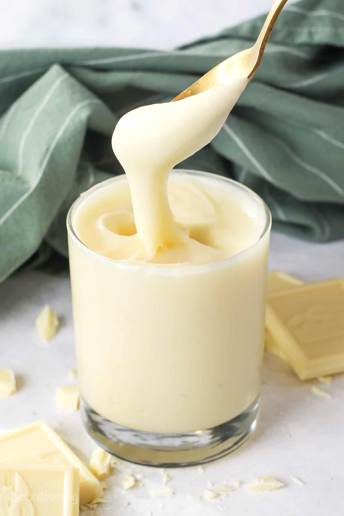 A spoon dipped into a glass of white chocolate ganache, next to scattered white chocolate pieces.