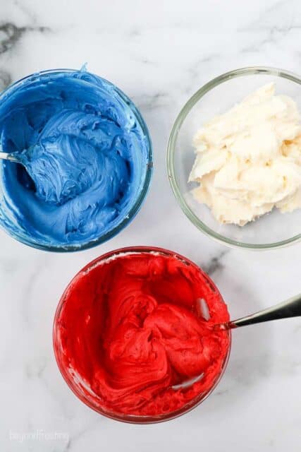 Overhead view of three bowls of frosting dyed blue, red and white