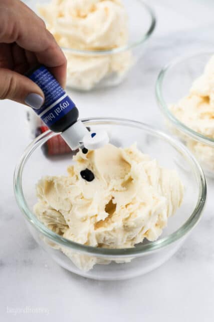 a hand squeezing a bottle of blue food coloring into a bowl of frosting