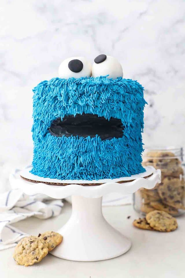 a white cake stand with a cookie monster cake and a jar of cookies in the background