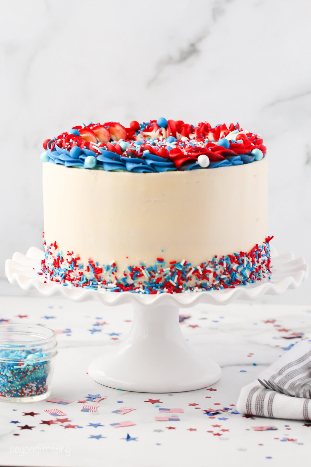 A frosted red white and blue layer cake decorated with piped red and blue frosting and a skirt of sprinkles on a cake stand.