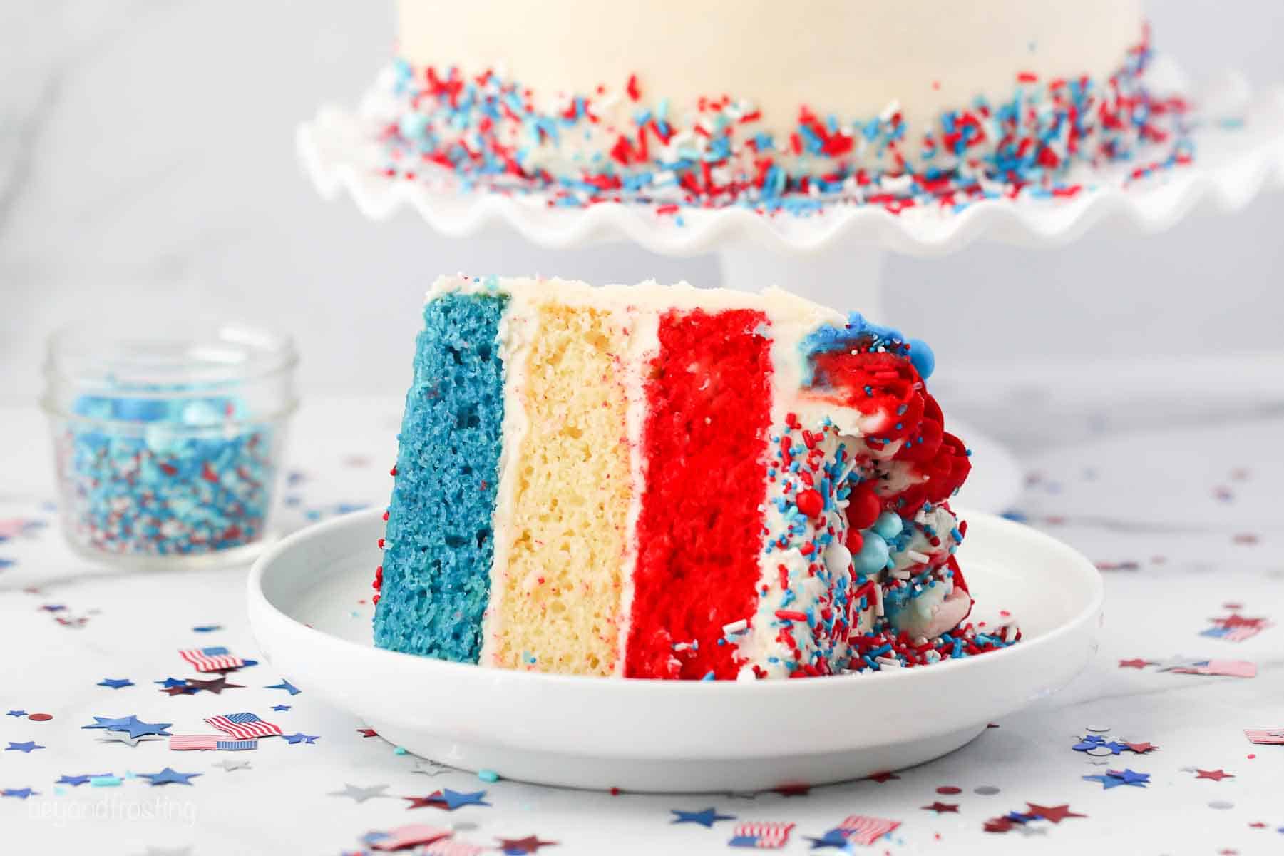 a slice of 3 layer cake with layers of blue, white and red cake. Patriotic confetti on the table.
