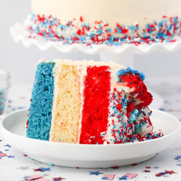 A slice of frosted red, white and blue layer cake on its side on a plate, with the rest of the cake on a cake stand in the background.