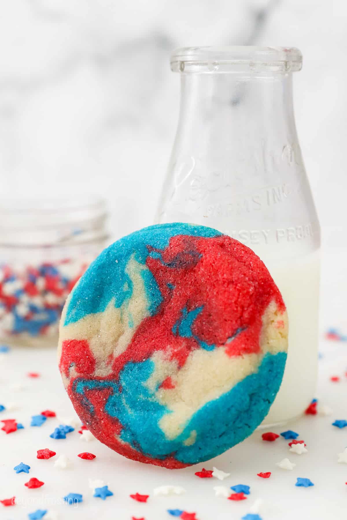 A patriotic sugar cookie propped up against a jug of milk surrounded by red, white, and blue confetti.
