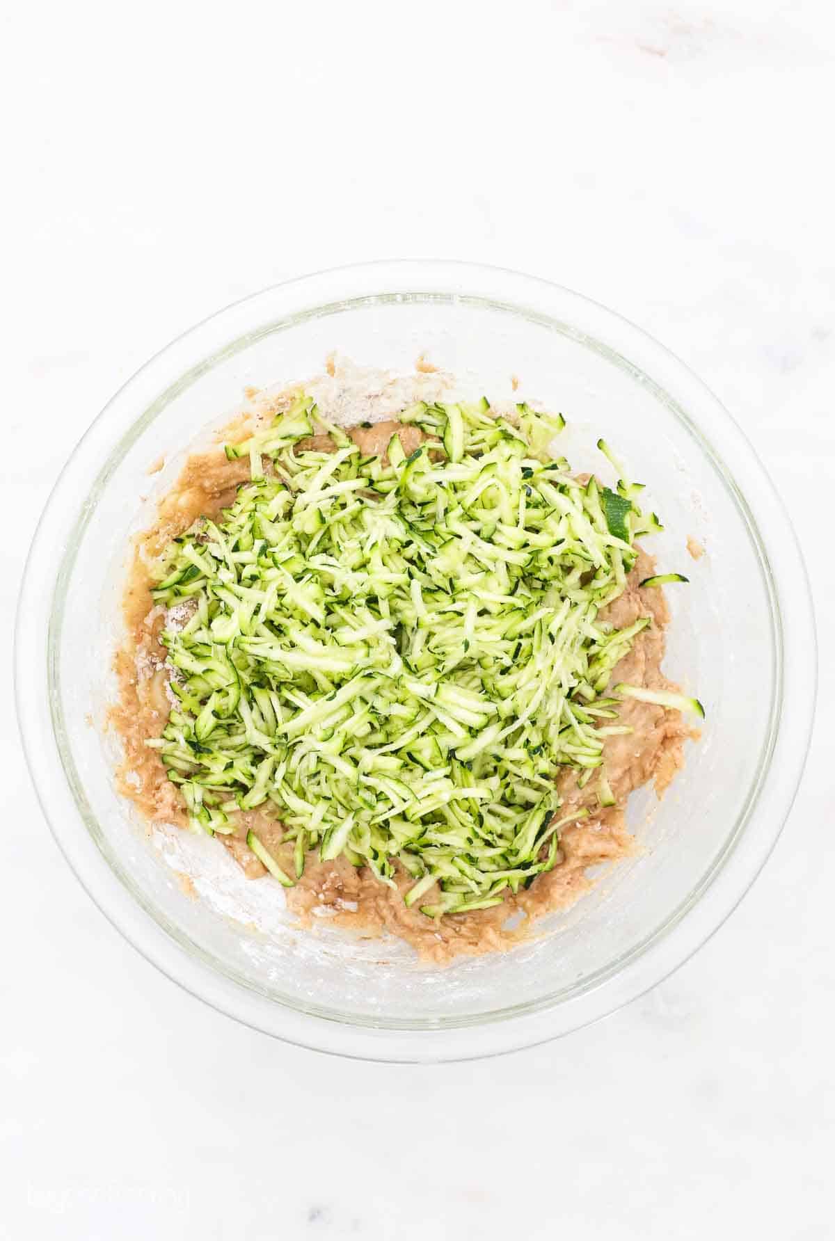 Shredded zucchini added to zucchini bread batter with cinnamon and vanilla in a mixing bowl.