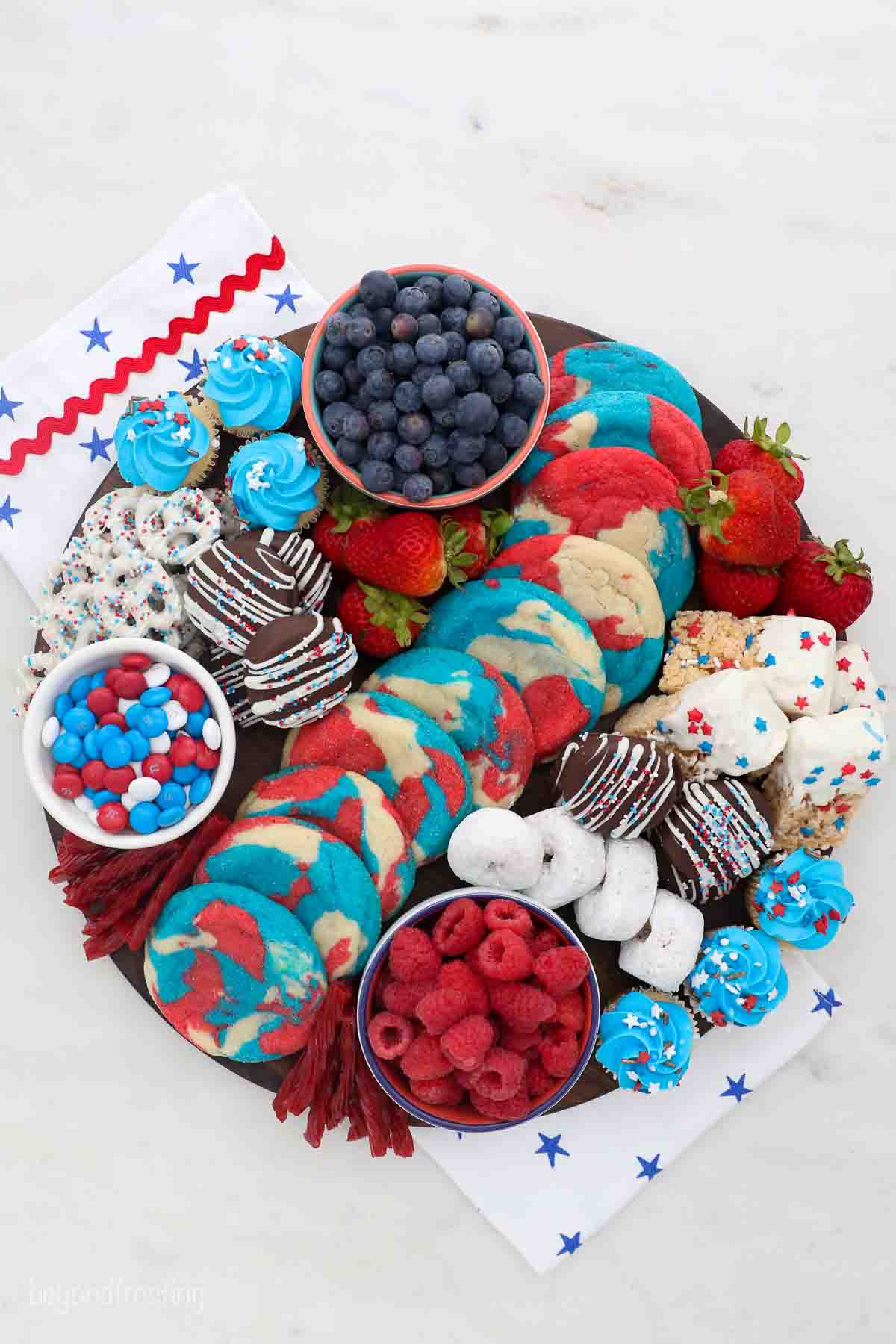 a round wooden board filled with red, white and blue themed desserts like cookies, candies and donuts