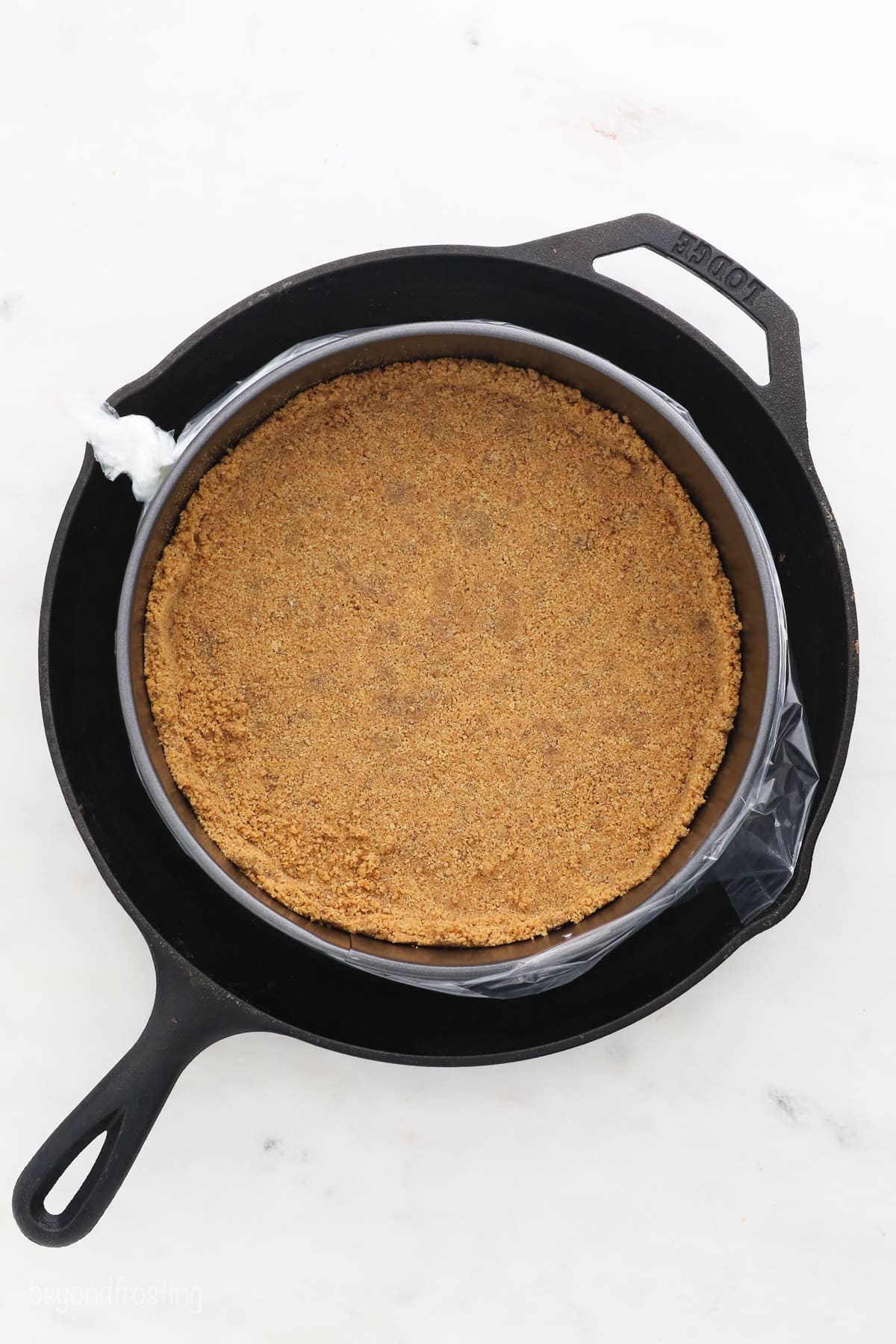Overhead view of a prepared graham cracker crust in a springform pan, placed in a water bath inside a skillet.