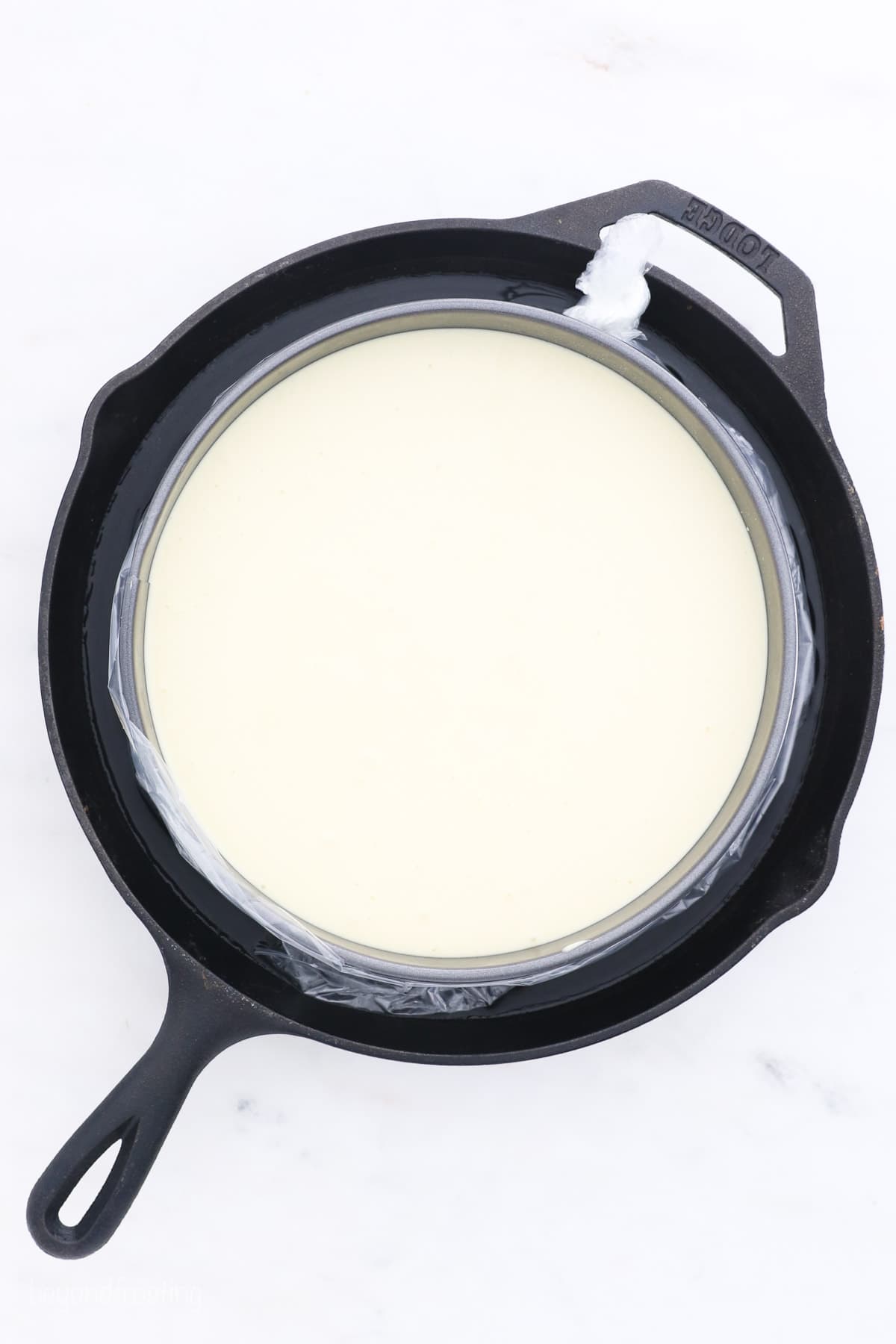 Overhead view of a prepared cheesecake in a springform pan, placed in a water bath inside a skillet.