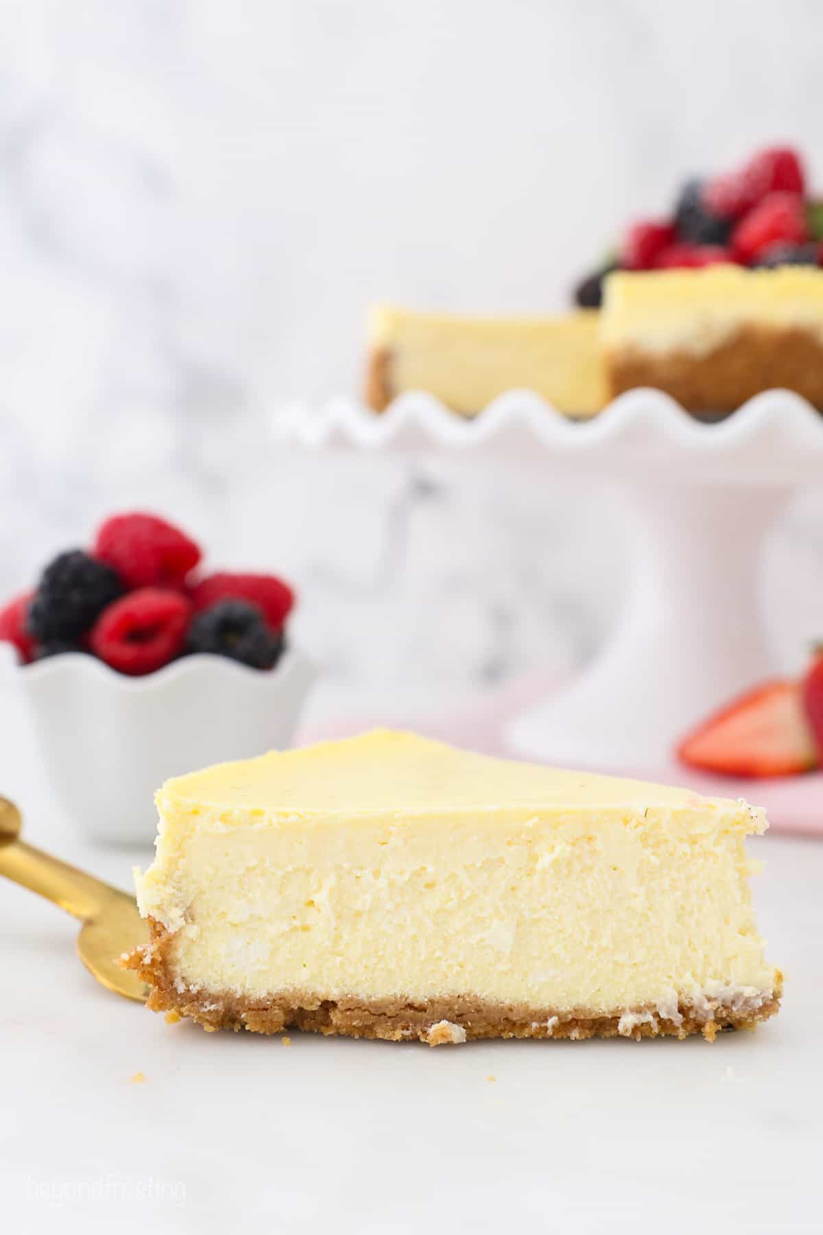 A slice of homemade cheesecake on a gold cake server, with the rest of the cheesecake on a cake stand in the background.
