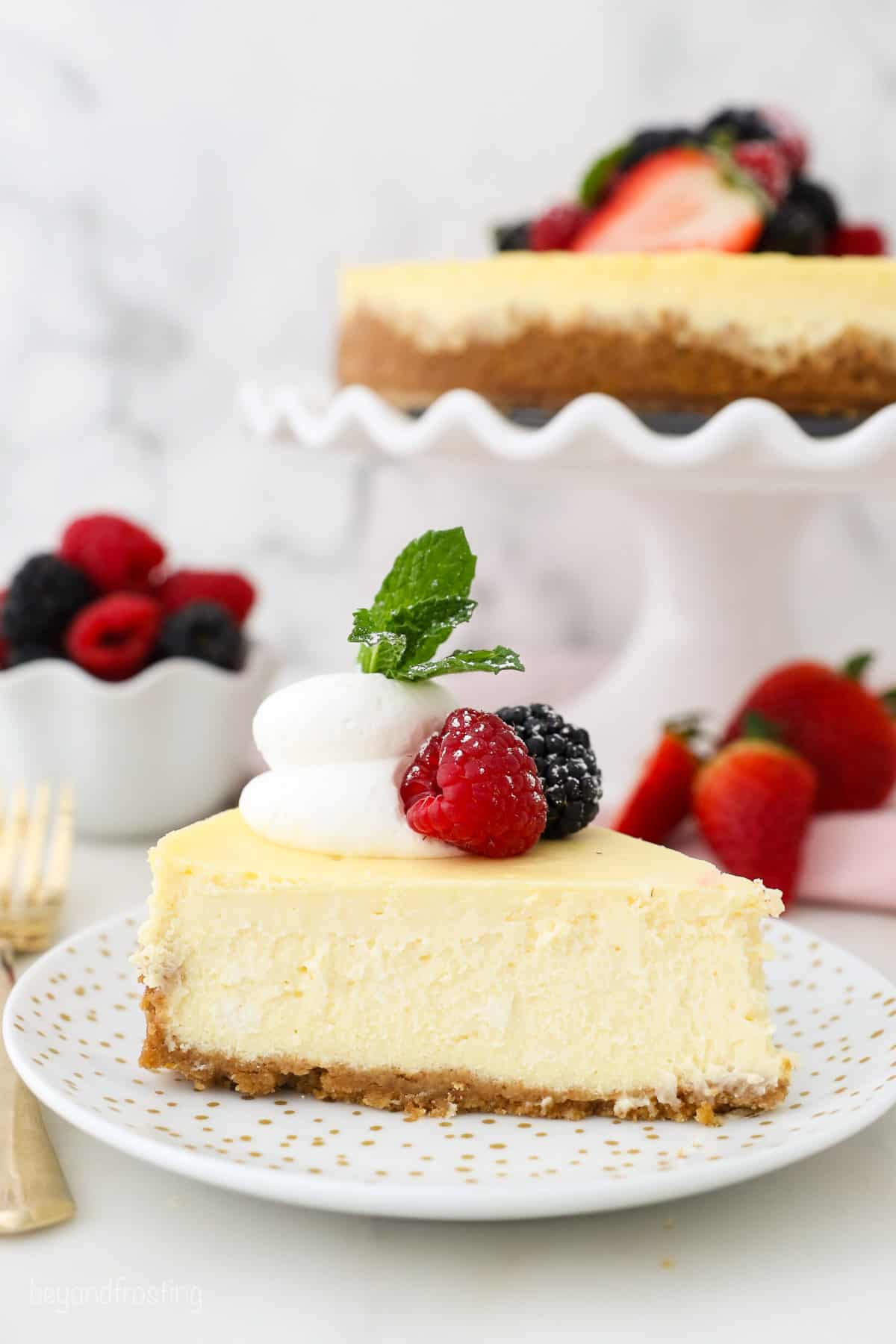 A slice of creamy cheesecake garnished with fresh berries and a swirl of whipped cream on a white plate, with the rest of the cheesecake on a cake stand in the background.