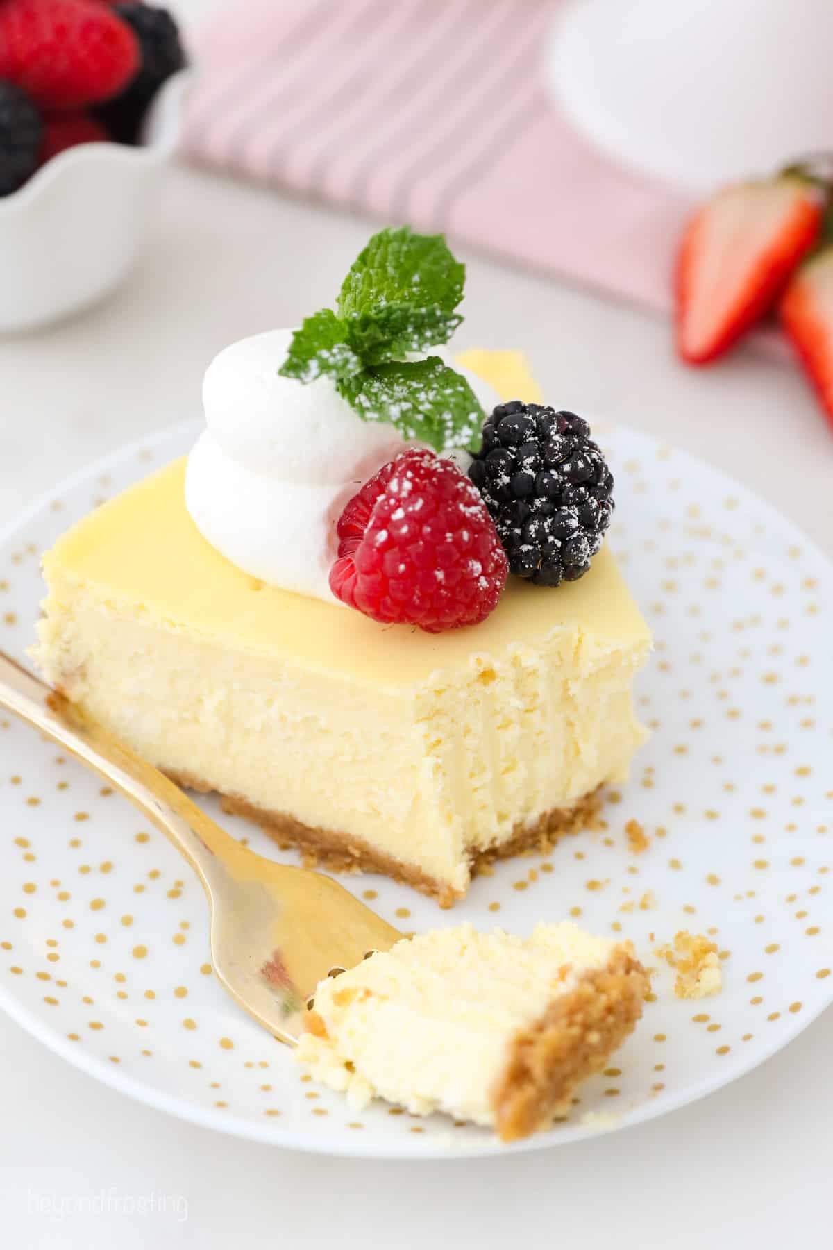 A forkful of cheesecake next to a slice of creamy cheesecake garnished with fresh berries and a swirl of whipped cream on a white plate.
