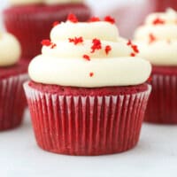 A red velvet cupcake topped with a swirl of cream cheese frosting and red sprinkles, with more cupcakes in the background.