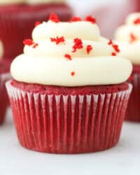 A red velvet cupcake topped with a swirl of cream cheese frosting and red sprinkles, with more cupcakes in the background.
