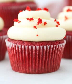 A red velvet cupcake topped with a swirl of cream cheese frosting and red sprinkles, with more cupcakes in the background.
