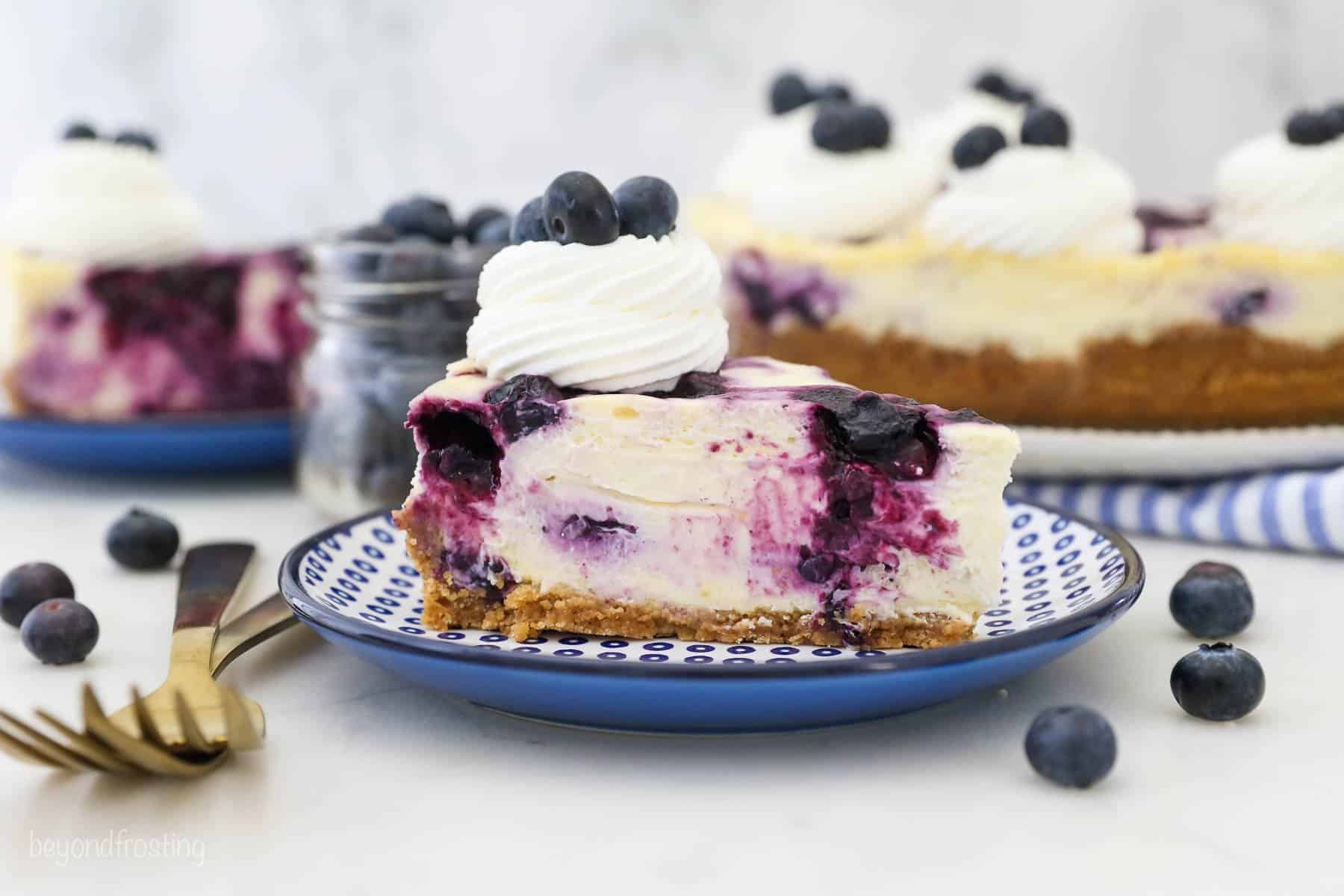 A blue polka dot plate surrounded by blueberries with a slice of blueberry cheesecake on the plate