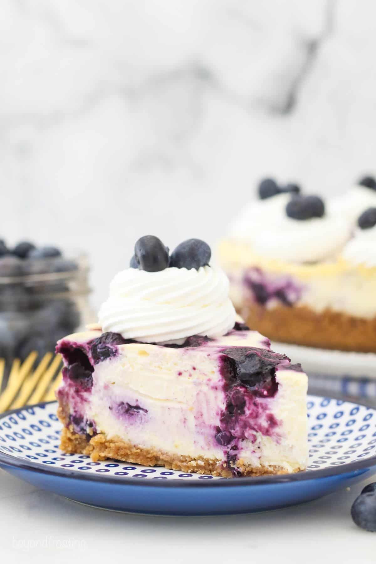 A slice of blueberry cheesecake topped with whipped cream and fresh berries on a plate with the rest of the cheesecake in the background.
