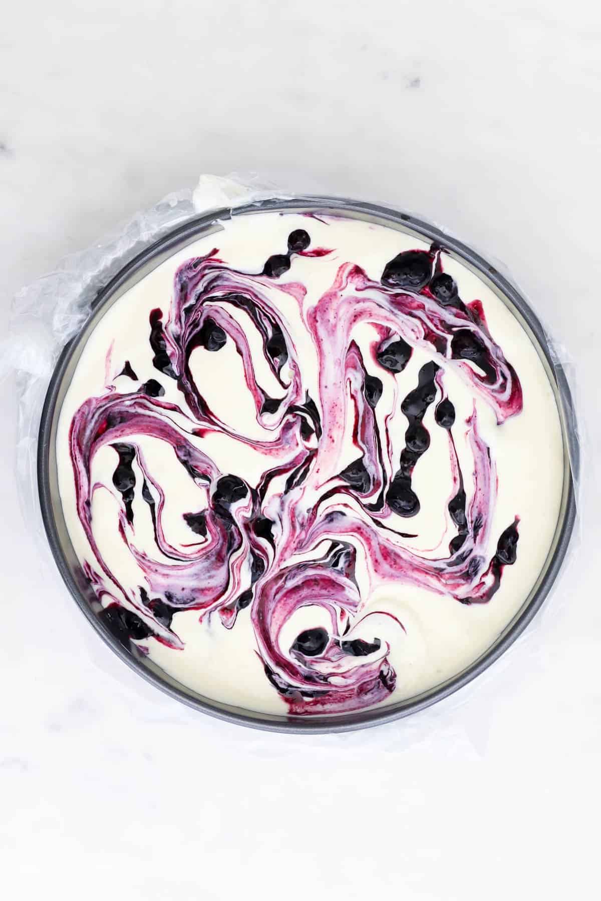 Overhead view of an unbaked cheesecake swirled with blueberry sauce inside a springform pan.