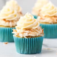 A coconut cupcake in a blue cupcake liner topped with a swirl of coconut frosting and toasted coconut, with more cupcakes in the background.