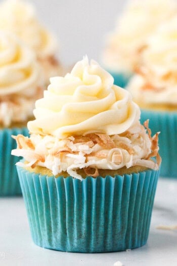 A coconut cupcake in a blue cupcake liner topped with a swirl of coconut frosting and toasted coconut, with more cupcakes in the background.