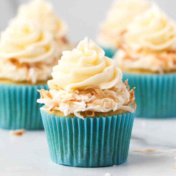 A coconut cupcake in a blue cupcake liner topped with a swirl of coconut frosting and toasted coconut, with more cupcakes in the background.