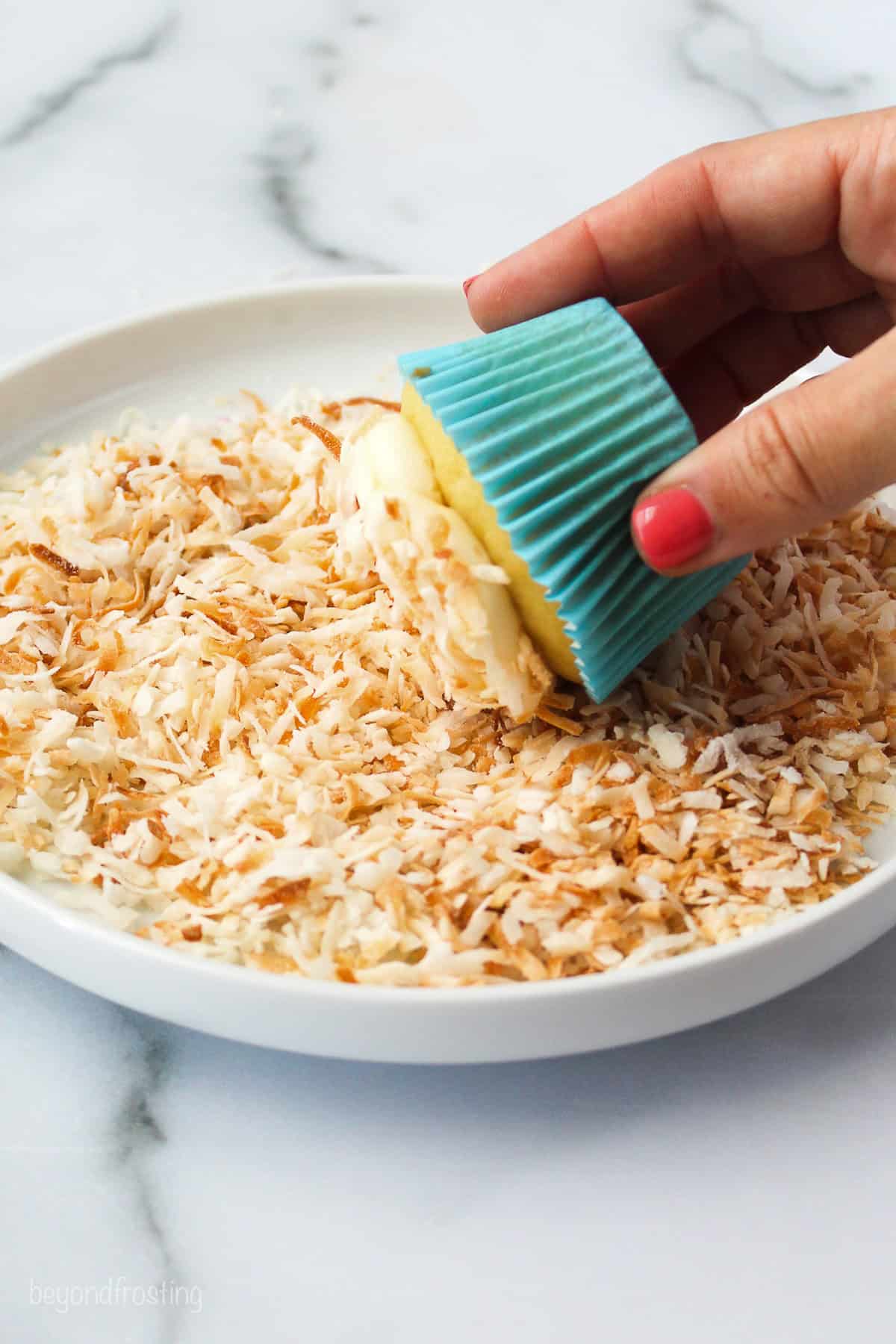 A hand rolling the top of a frosted cupcake into a shallow dish of toasted coconut.