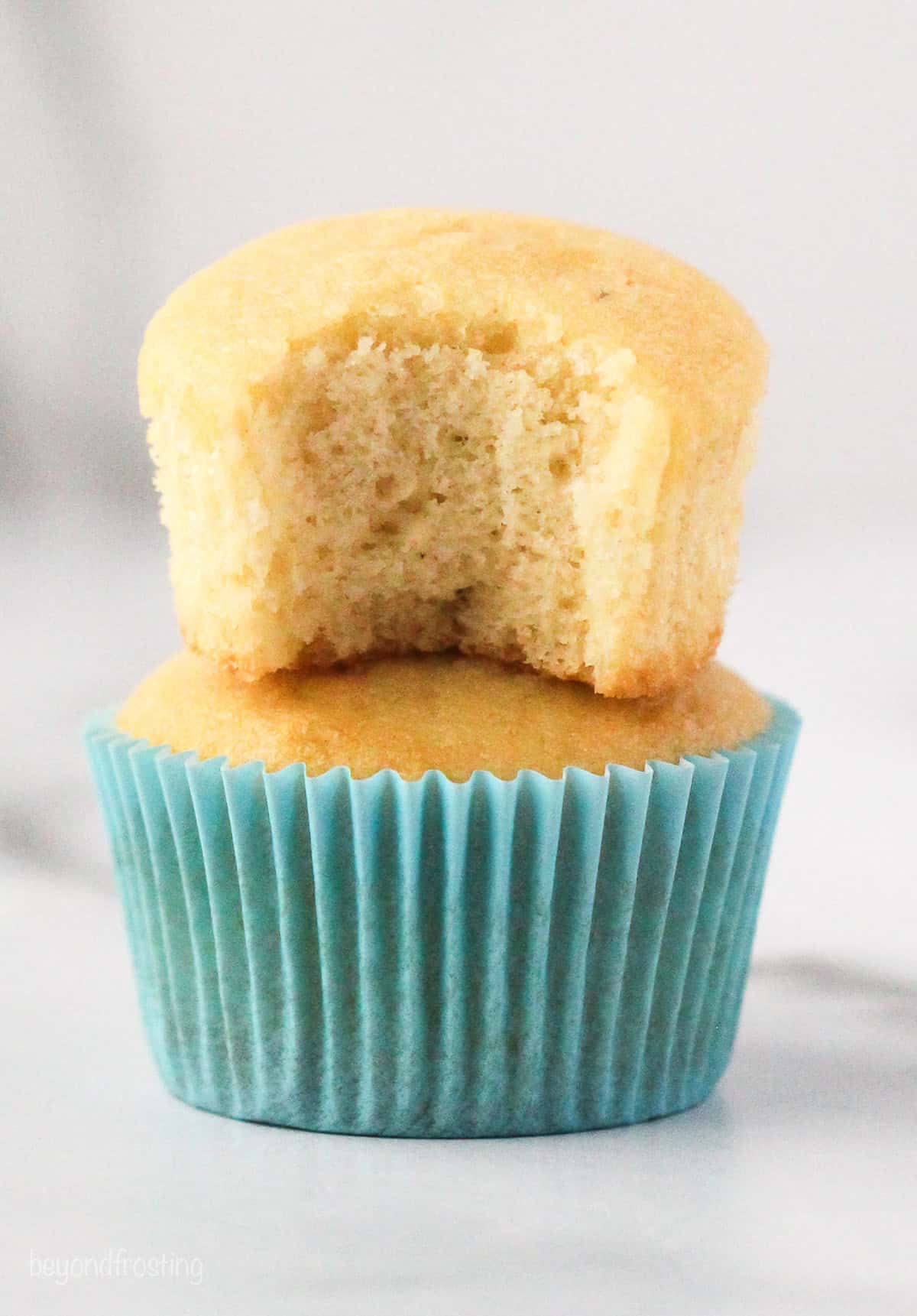 A coconut cupcake with a bite missing stacked on top of a second cupcake in a blue cupcake liner.