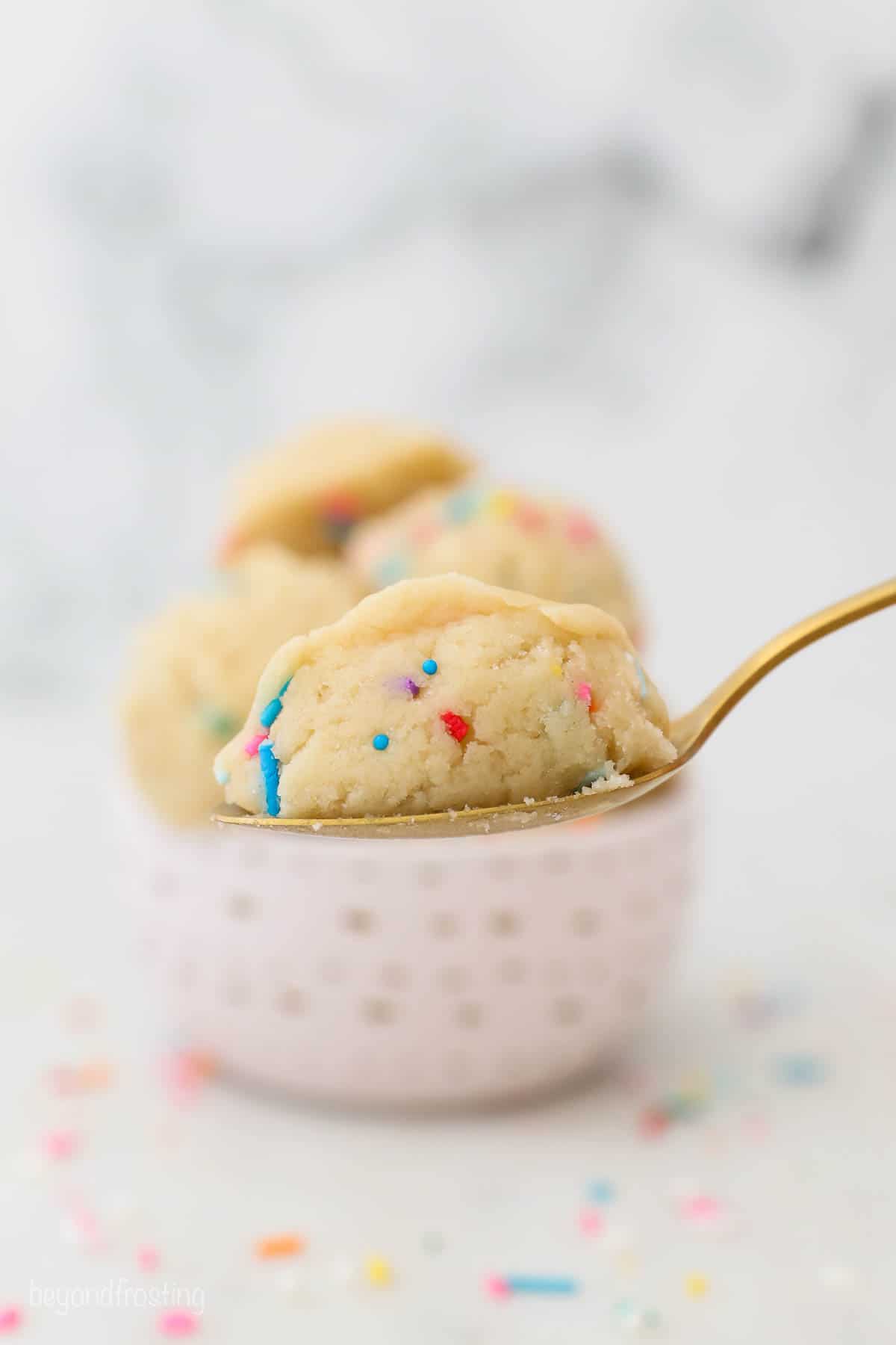 Sweet, buttery edible sugar cookie dough is egg-free and made with heat-treated flour so it's safe to eat straight from the mixing bowl. Close up of a gold spoon with a scoop of edible sugar cookie dough, with more cookie dough scoops in a bowl in the background.
