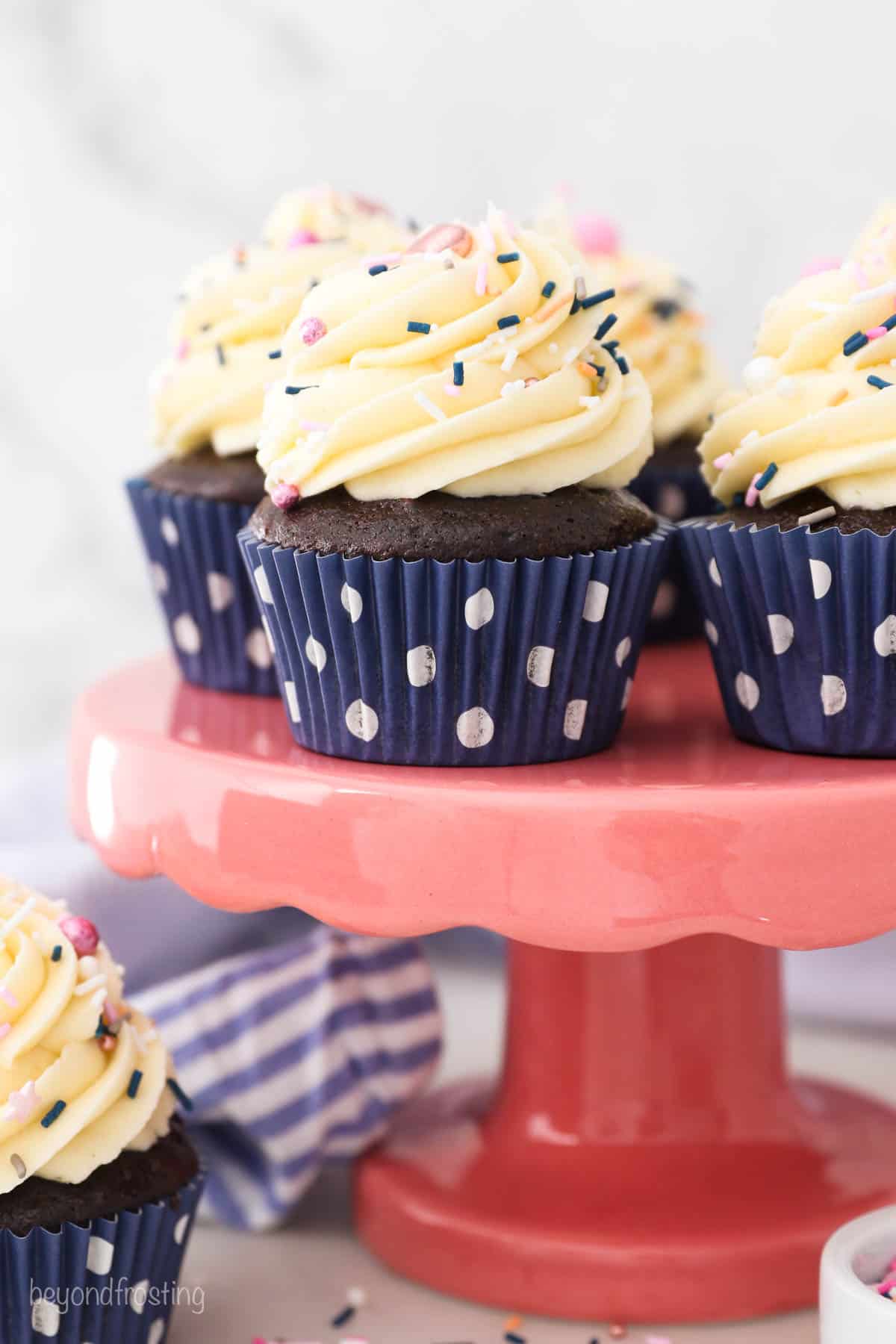 A pink cake stand with chocolate frosted cupcakes in navy blue polka dot wrappers