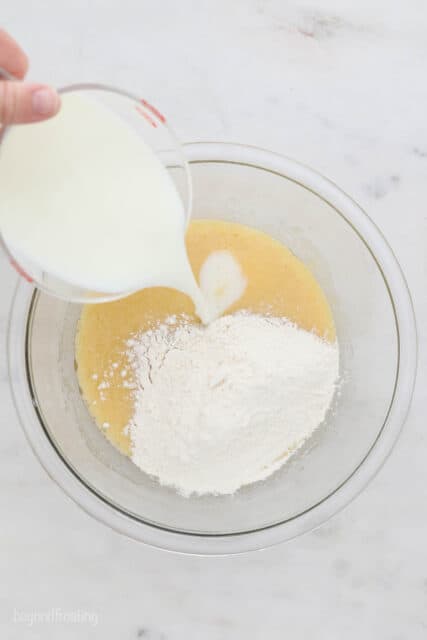 a glass mixing bowl with cake batter, flour and milk being poured in
