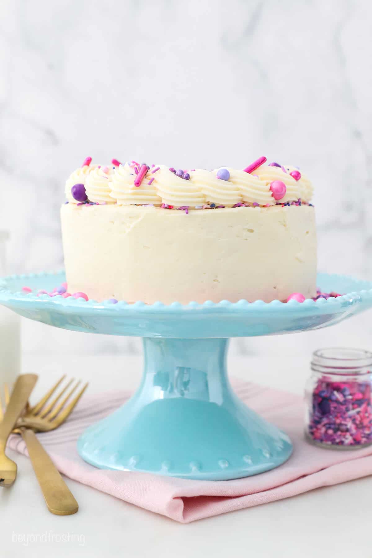 a decorated vanilla cake on a teal cake stand, pink napkin and gold forks