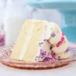 a small slice of vanilla cake with sprinkles on a pink glass plate with a cake stand in the background