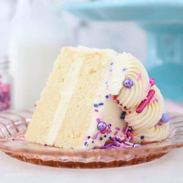 a small slice of vanilla cake with sprinkles on a pink glass plate with a cake stand in the background