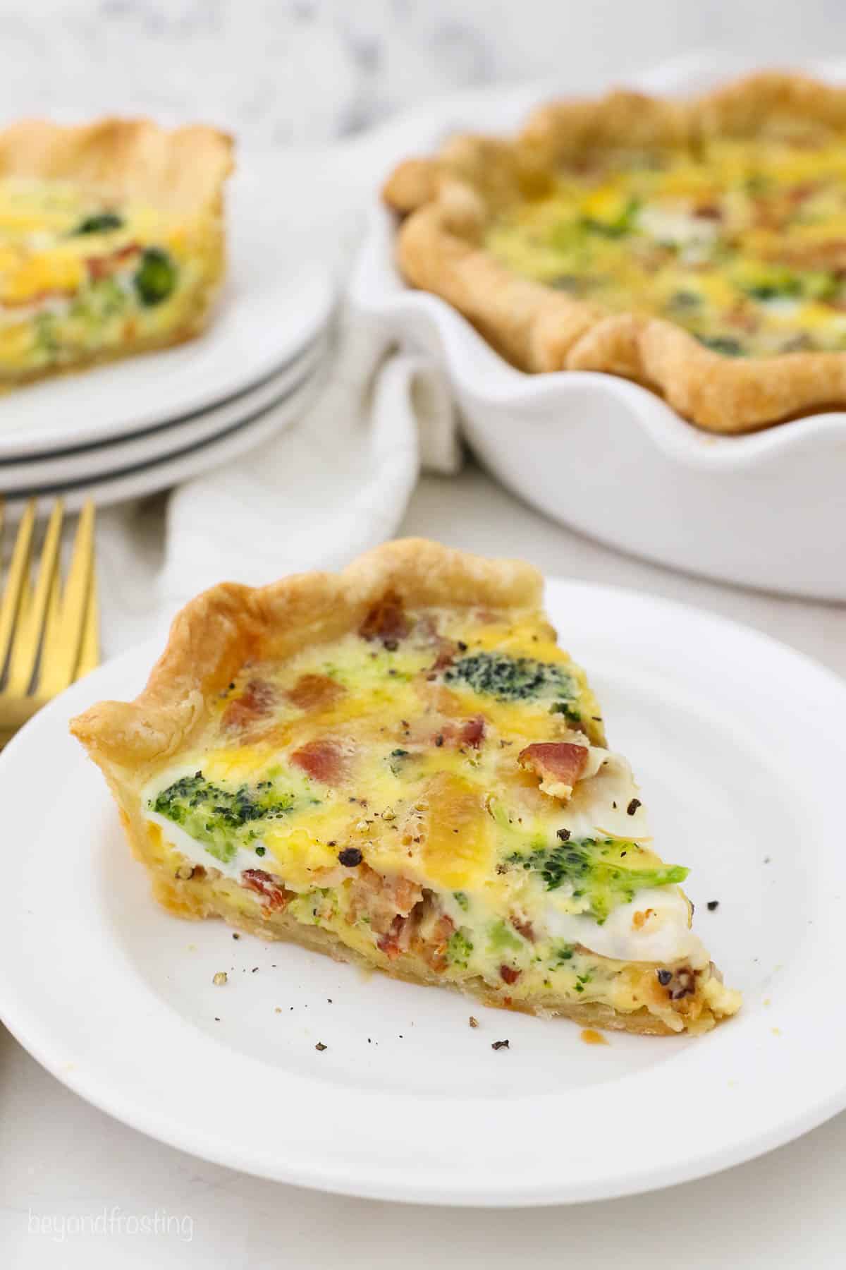 A slice of broccoli bacon quiche on a white plate with the rest of the quiche in the background