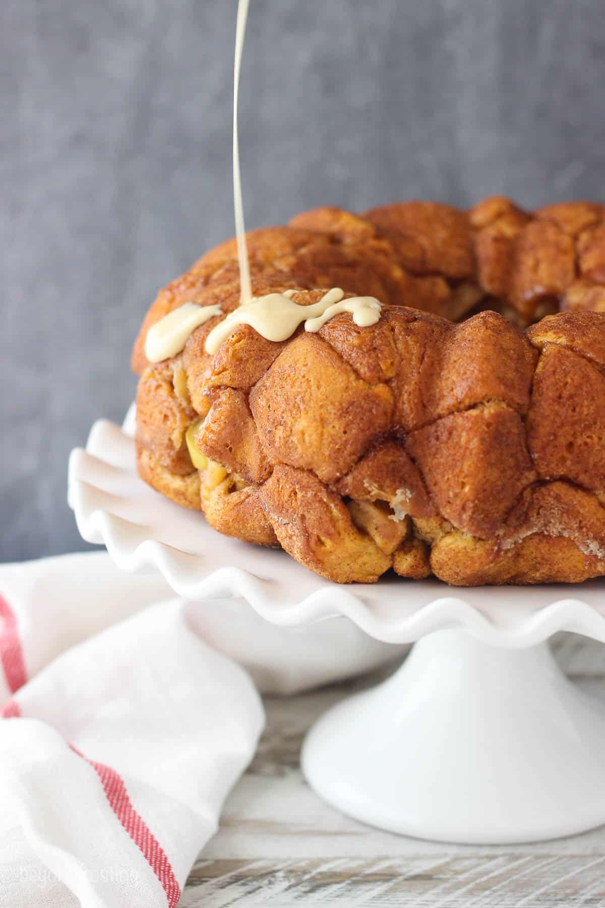 Apple pie pull apart bread on a cake stand with maple glaze being drizzled overtop