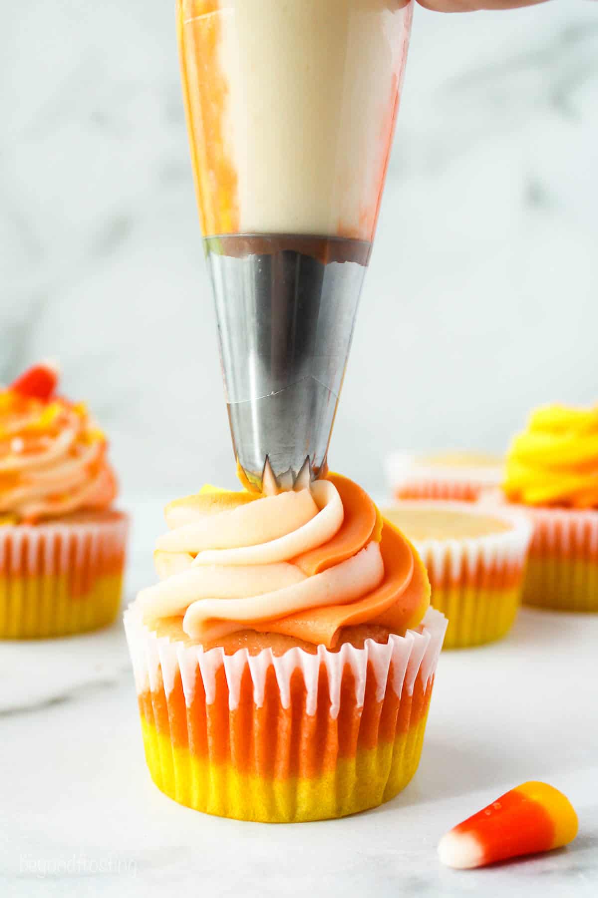 A piping bag frosting a cupcake with white, orange and yellow frosting