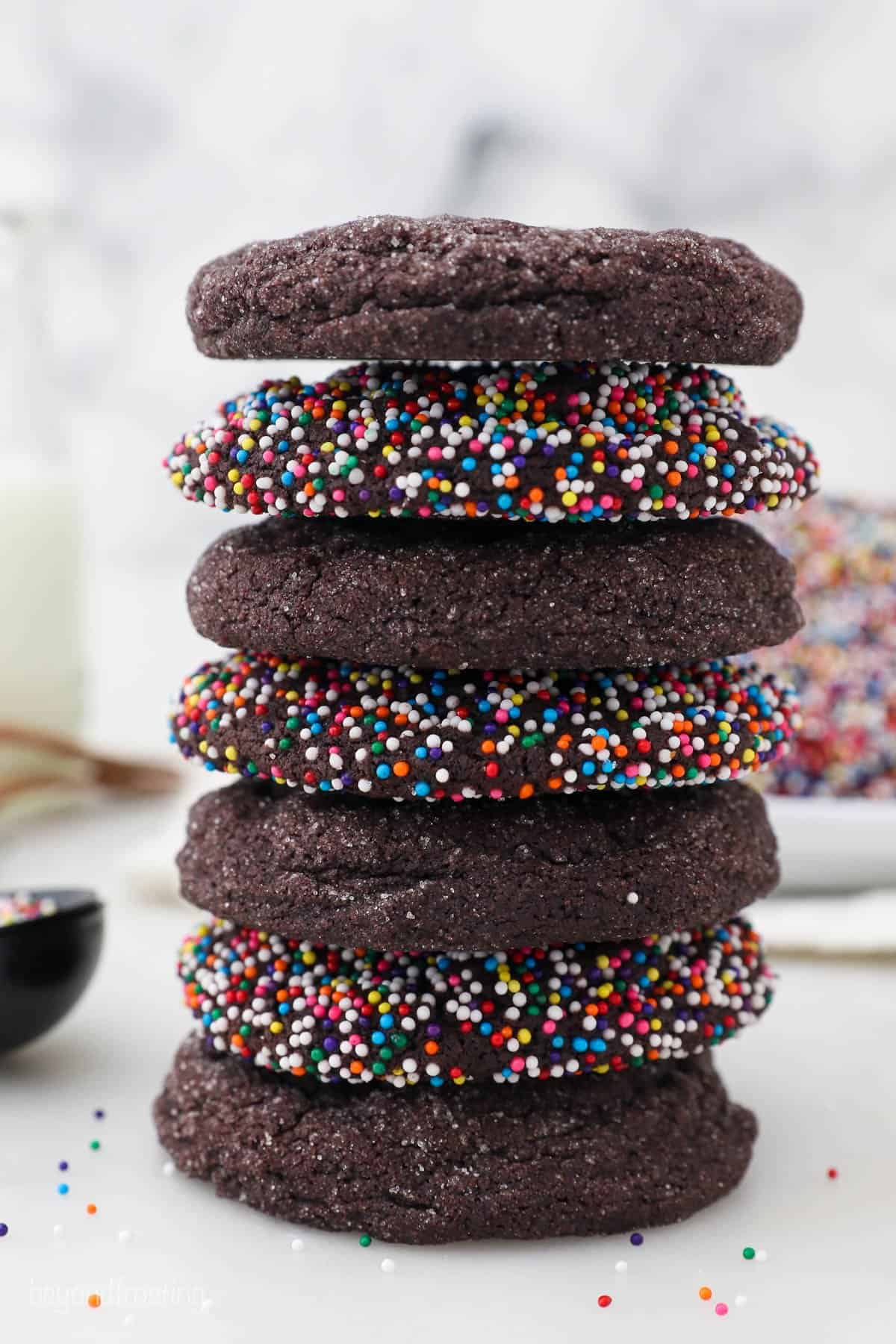 Seven chocolate sugar cookies stacked on top of one another with a bowl of rainbow sprinkles in the background