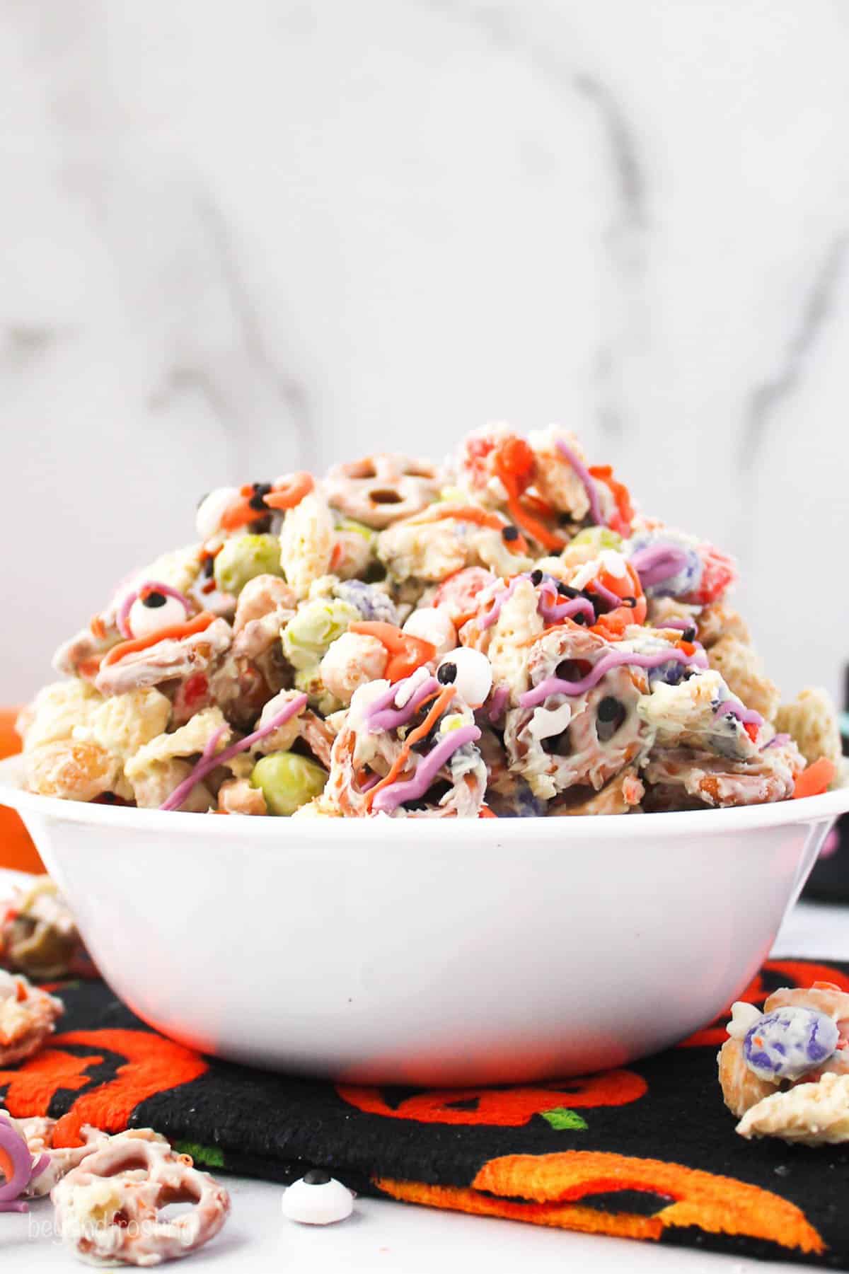 a white bowl sitting on a pumpkin towel. The bowl is filled with with a halloween snack mix