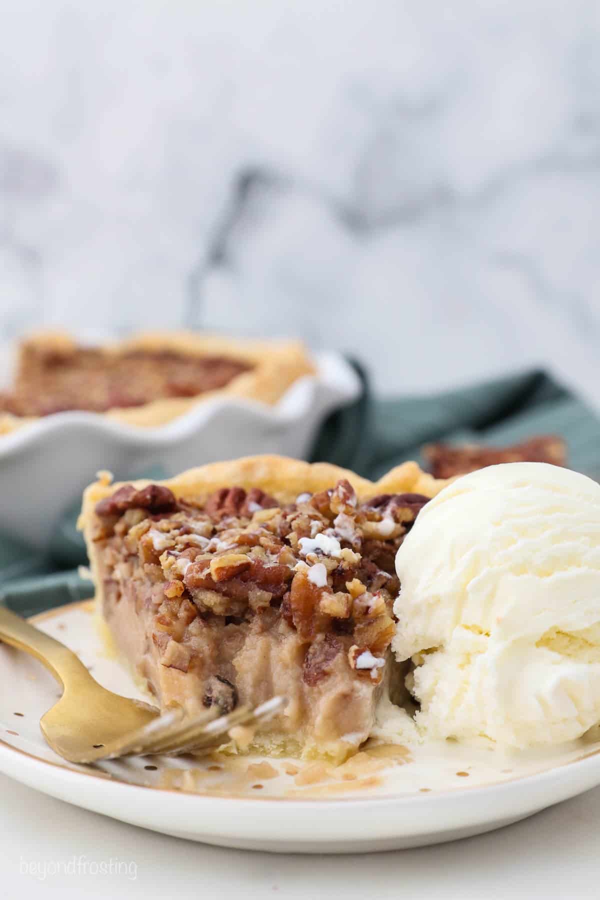 A piece of pecan pie on a white plate next to a fork and a scoop of vanilla ice cream