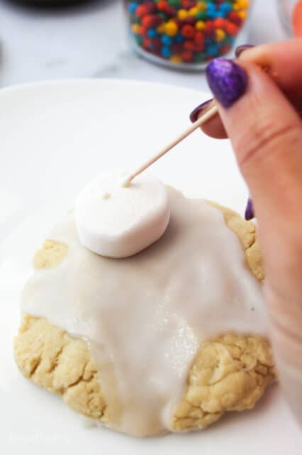 frosted sugar cookie topped with a marshmallow being dotted with white chocolate
