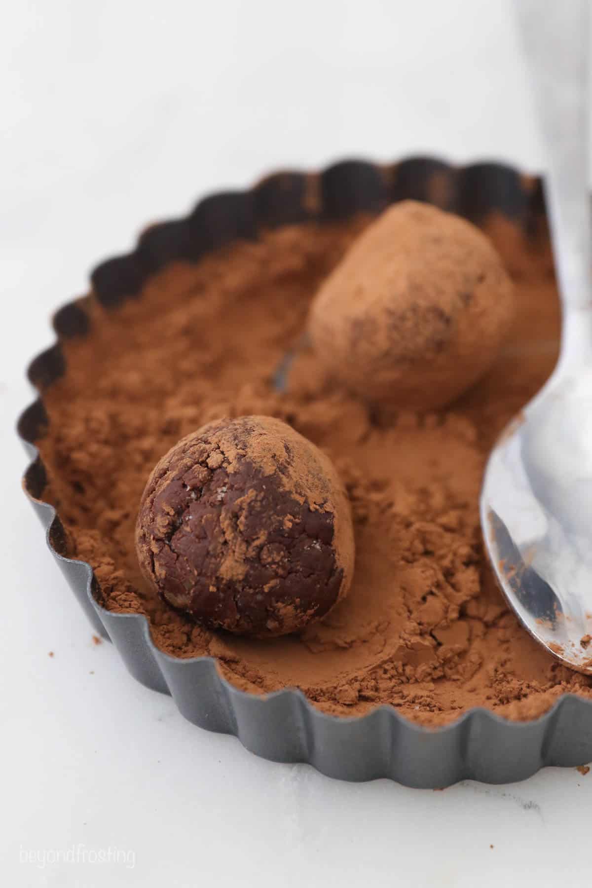 Two truffles covered with cocoa powder in varying degrees beside a metal spoon