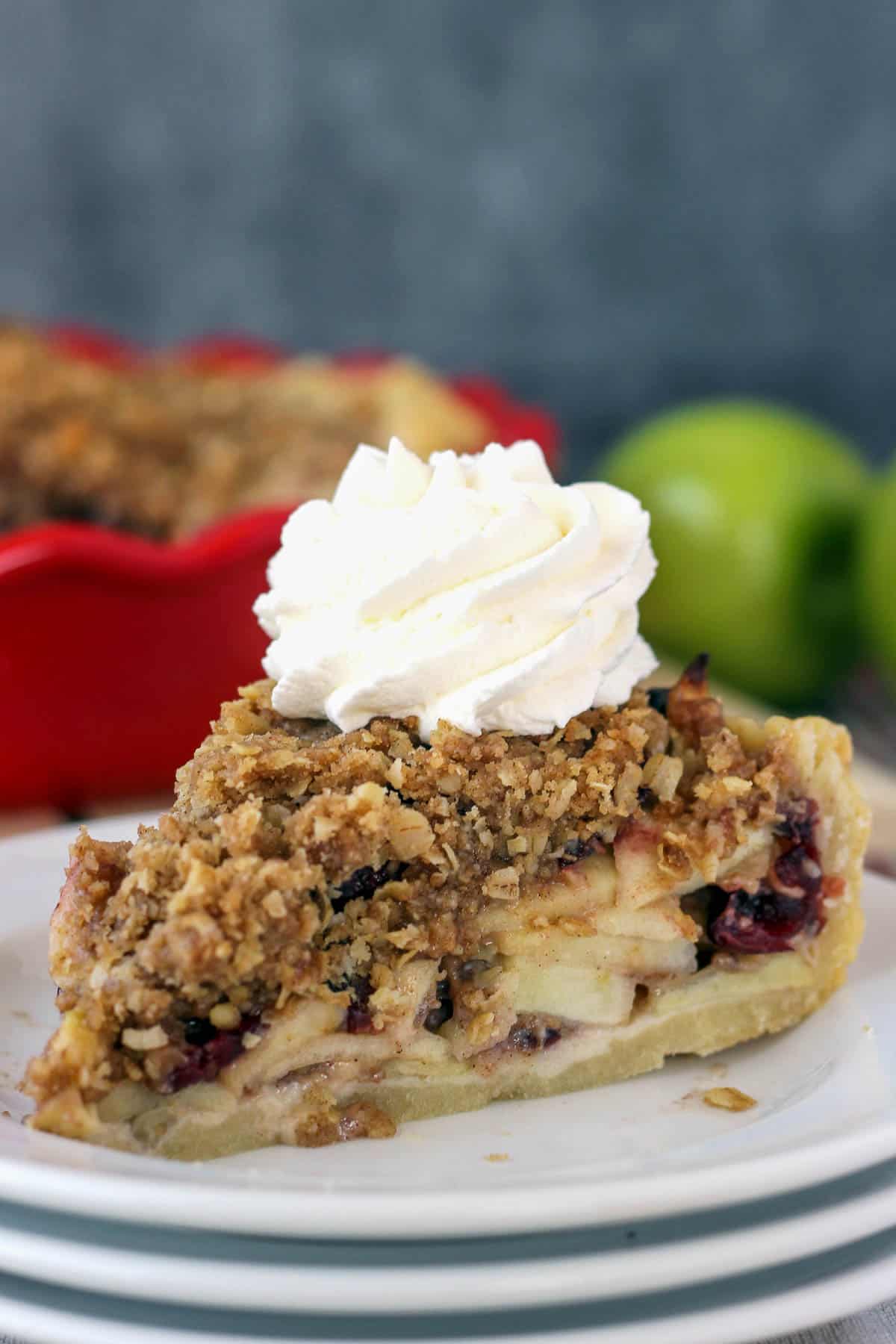 A slice of pie on a plate with whipped cream on top and fresh apples in the background