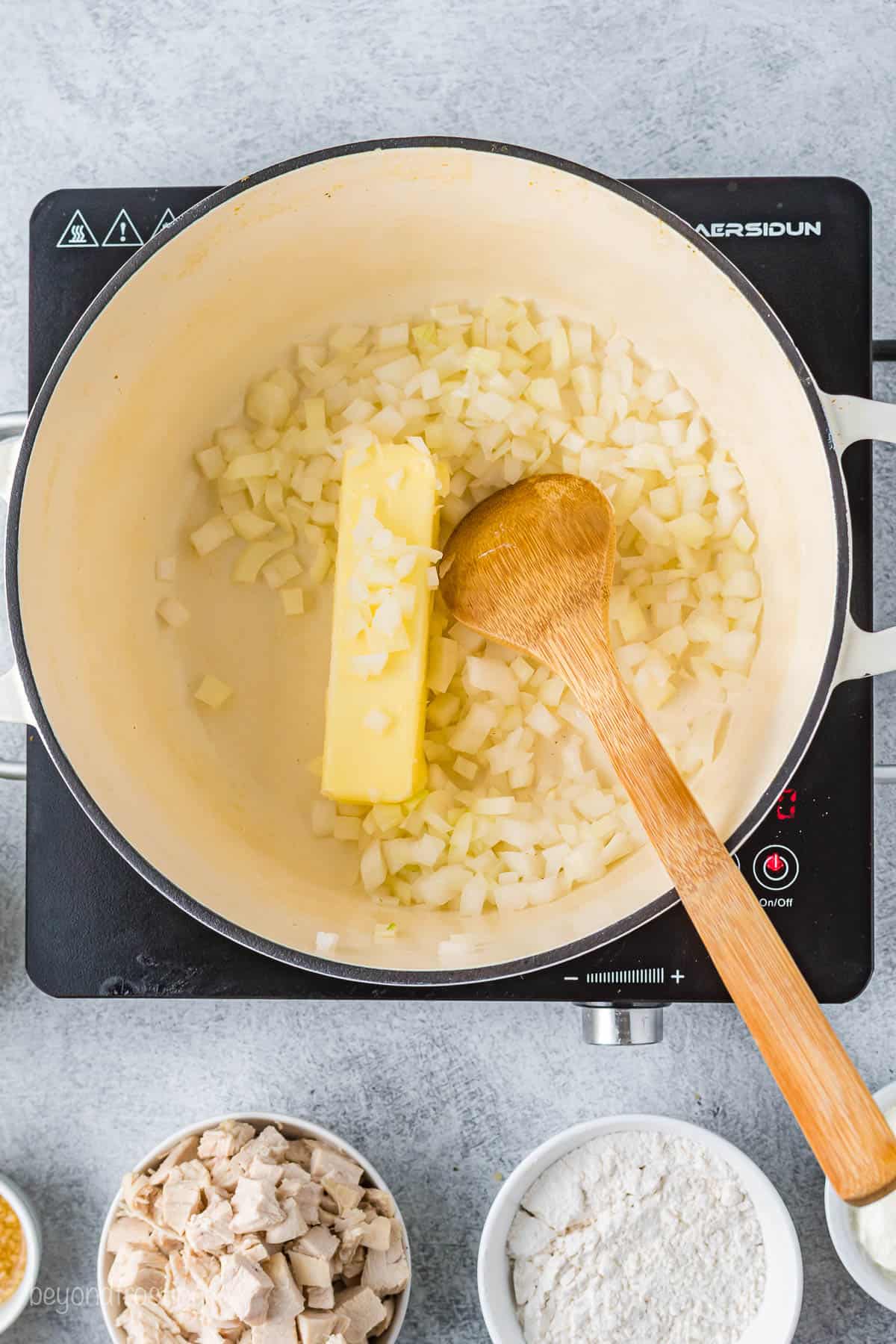 A stick of butter inside of a saucepan with chopped onion and a wooden spoon