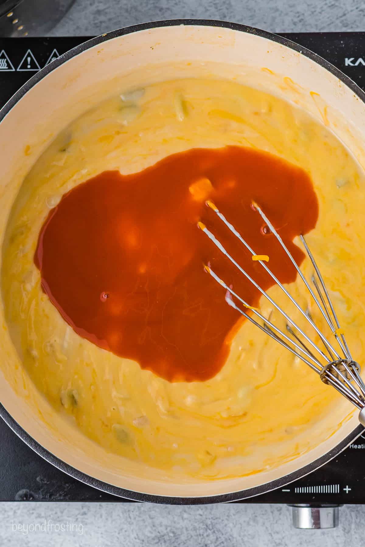 A whisk mixing Frank's hot sauce into a saucepan full of cheese sauce